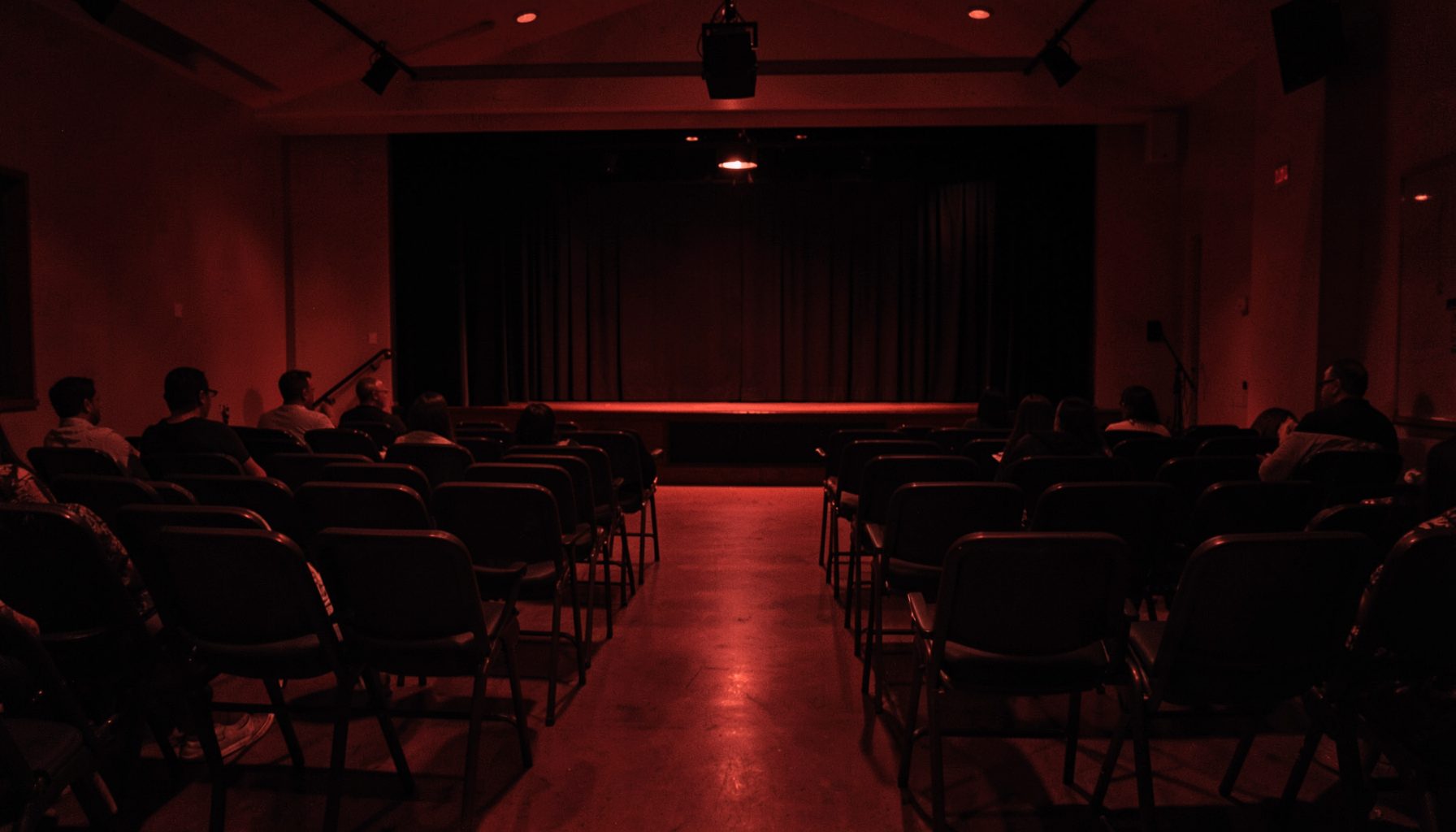 A warm, dimly lit community gathering space with rows of empty chairs facing a small stage, bathed in deep red and black tones, evoking a quiet sense of anticipation