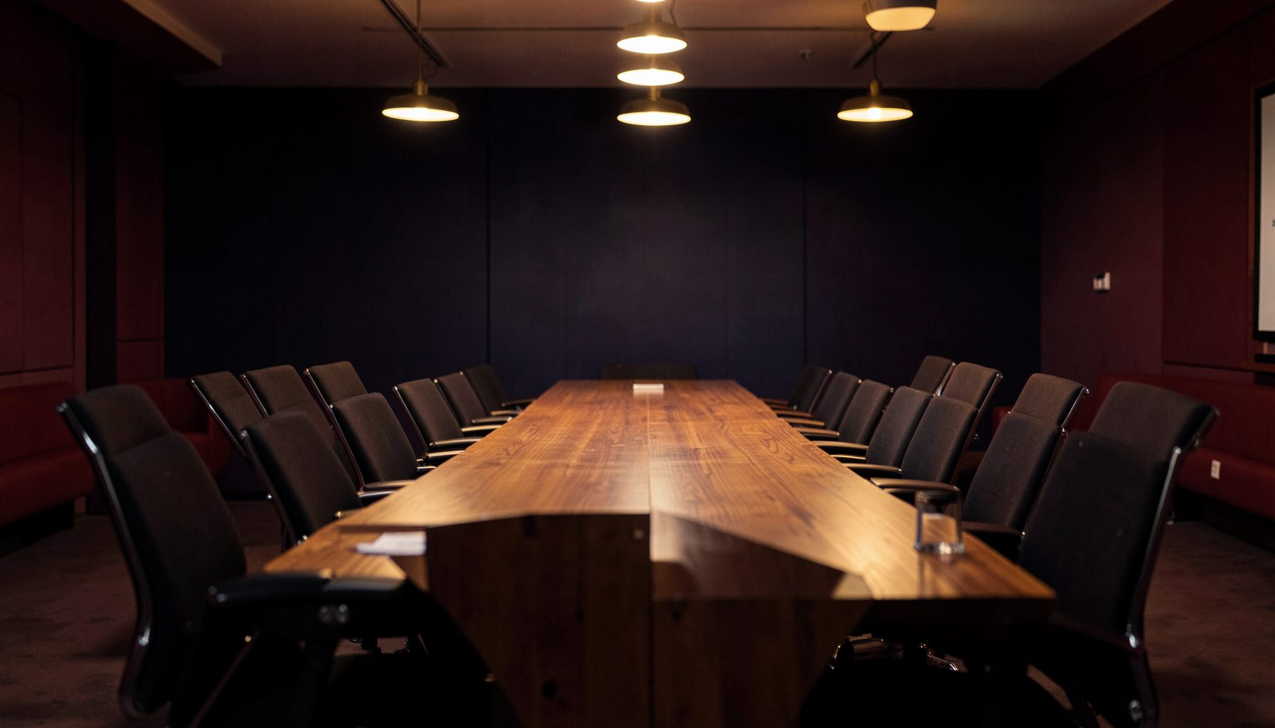 A warmly lit room with a long wooden table and empty chairs arranged for a panel discussion, deep navy and burgundy tones, soft amber glow from overhead fixtures