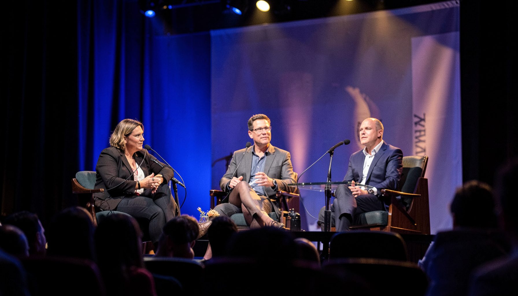 A panel discussion on stage with microphones and a banner, auditorium lighting in deep blue and warm gold tones.